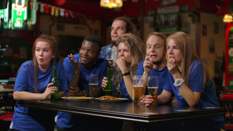 A-multi-ethnic-group-of-friends-of-fans-in-blue-t-shirts-will-watch-a-match-on-TV-in-a-bar-watch-a-tense-exciting-moment-and-celebrate-a-goal-scored.-Hugging-having-fun-and-making-a-glass-of-beer.